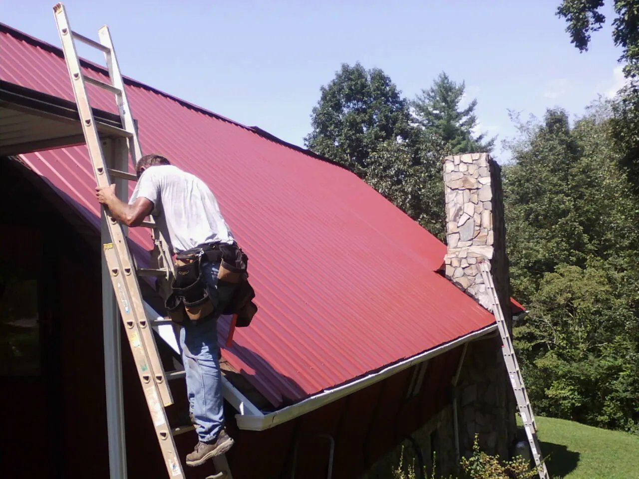 Roofer on a ladder beside a red metal roof and stone chimney — Soffit & Fascia Repair by Roofing Brothers in Blauvelt